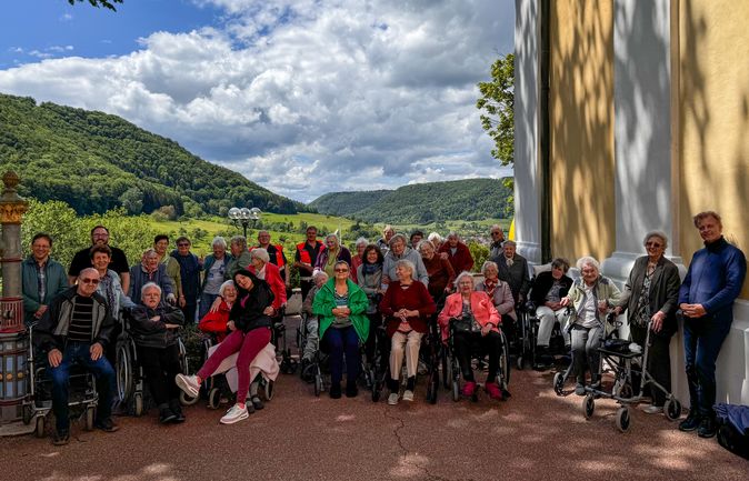 Seniorengruppe mit Rollstühlen vor gelber Klosterfassade und Hügellandschaft